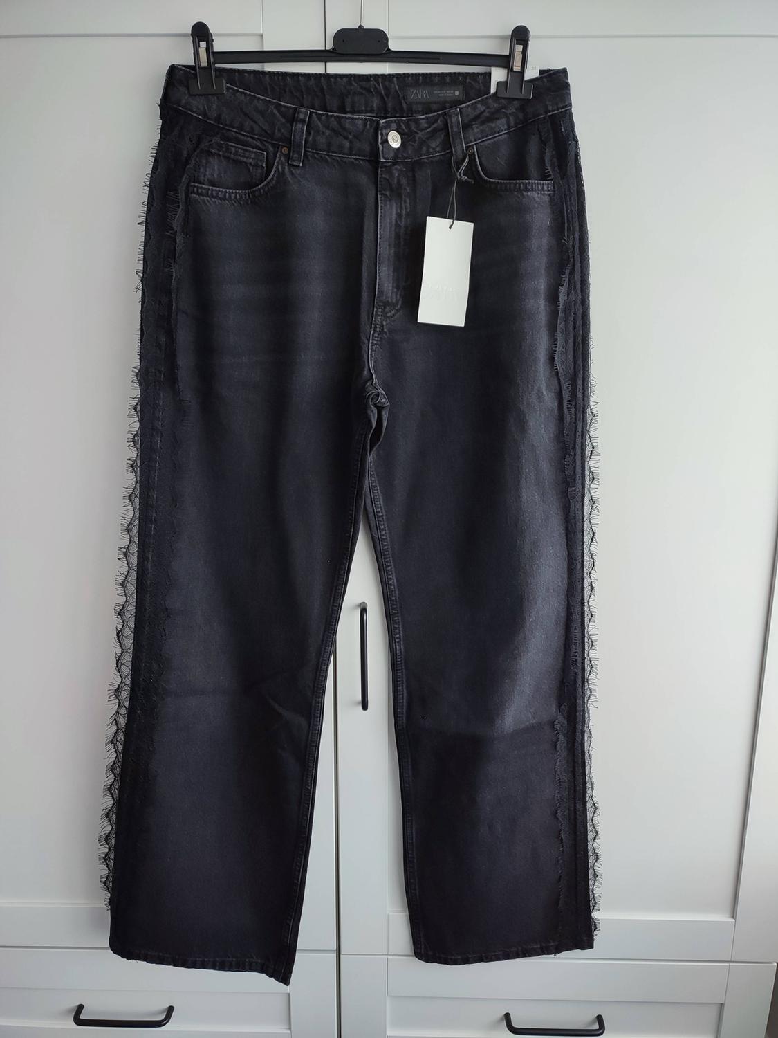 Black zara jeans with frayed detailing on the sides, hanging on a hanger in front of a white wardrobe.