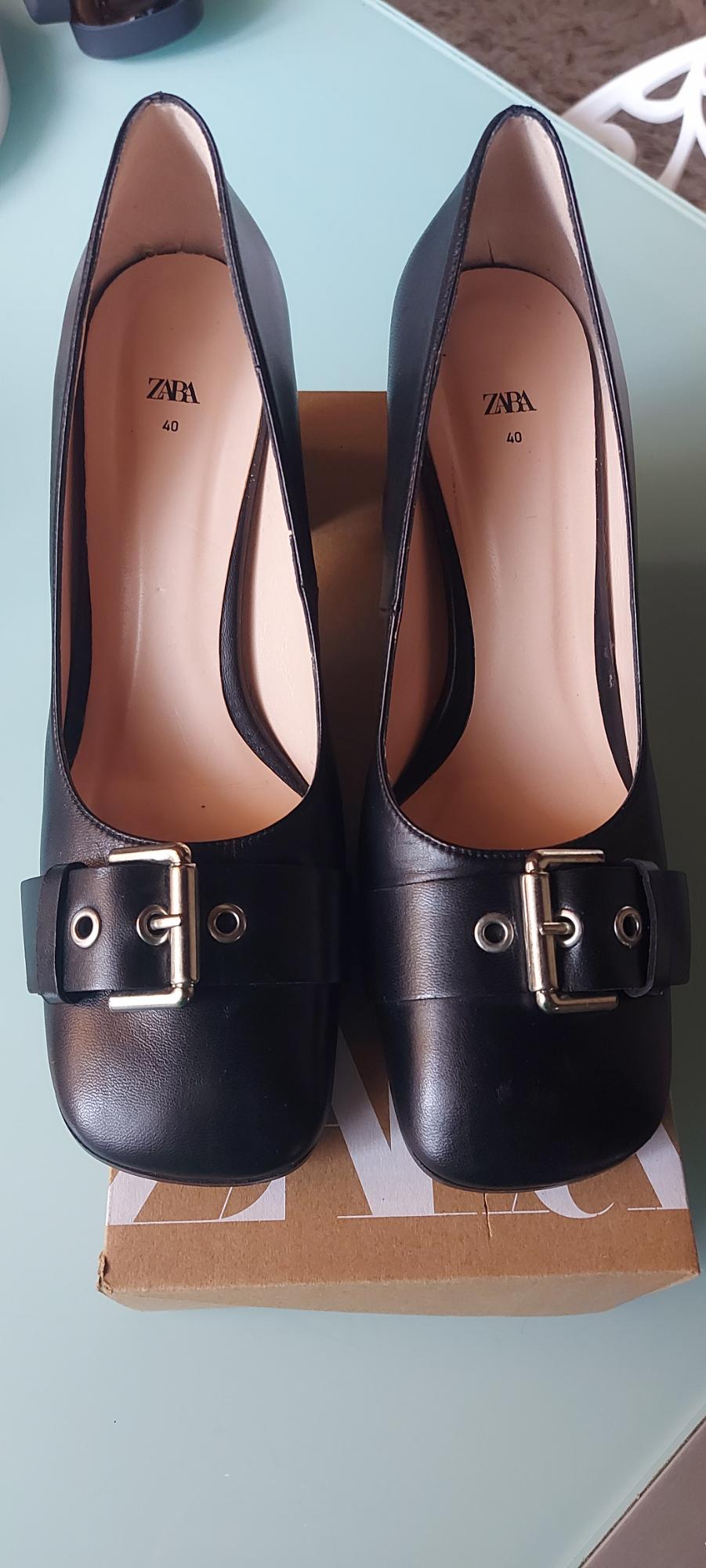 Black high-heeled shoes with a metallic buckle on a cardboard box. Beige interior with text "ZARA 40". Light blue background.