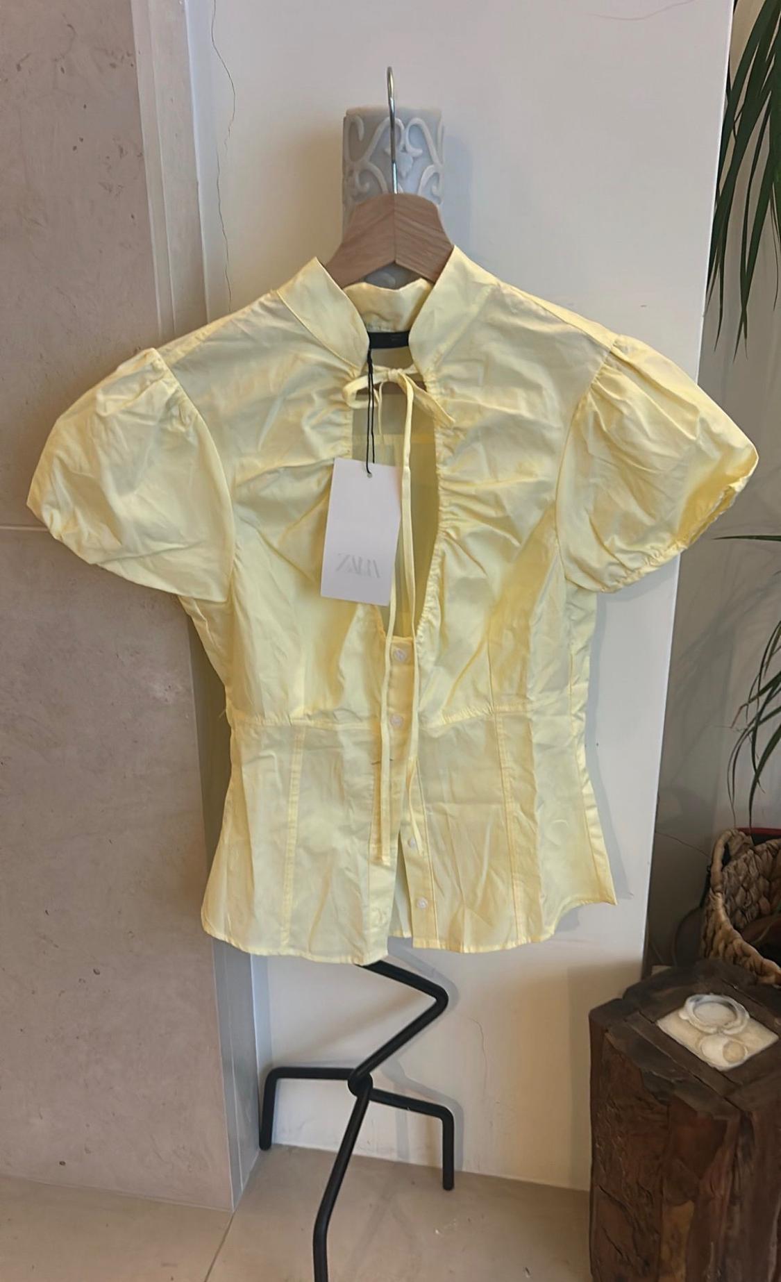 Yellow short-sleeved blouse with puffed sleeves and a neck tie, hanging on a hanger on a stand. Label with the text ZARA.