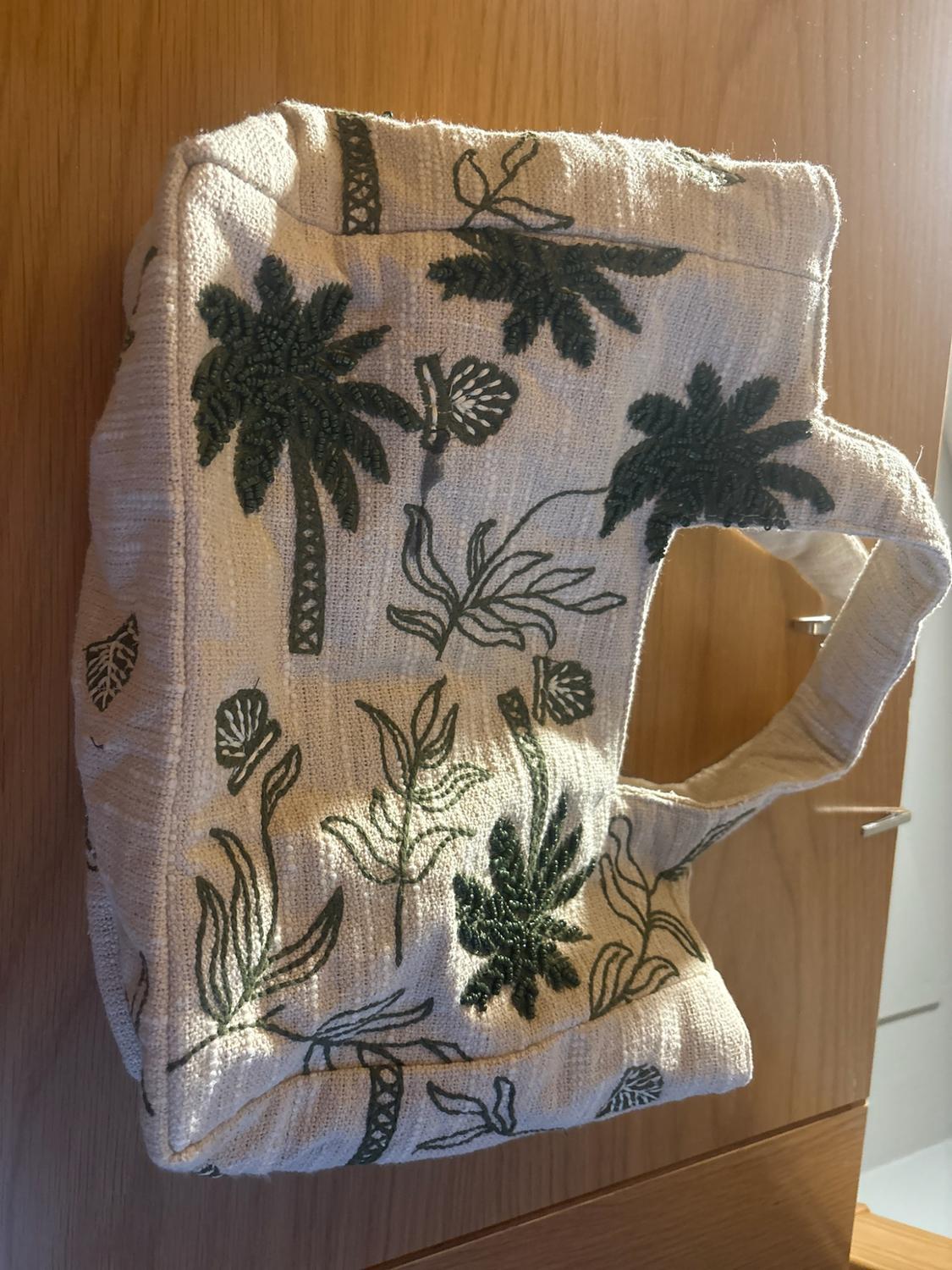 Beige fabric bag featuring a dark green palm tree and tropical leaf design, hanging on a wooden door.