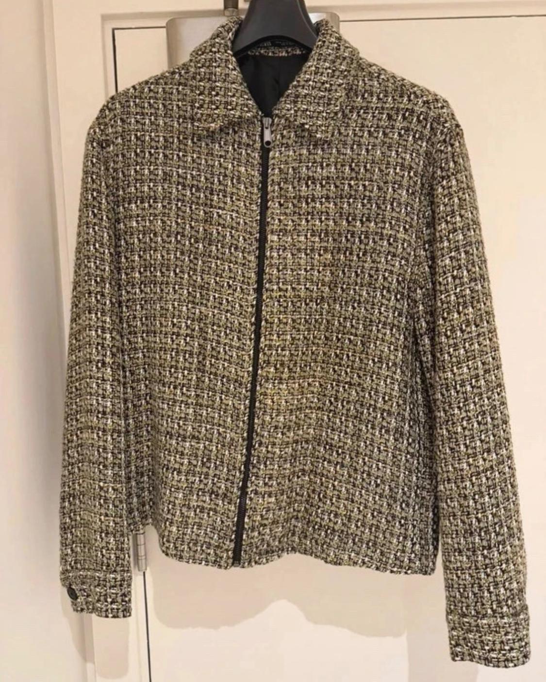 Textured fabric jacket in green and black tones with a front zip, hanging on a hanger in front of a white door.