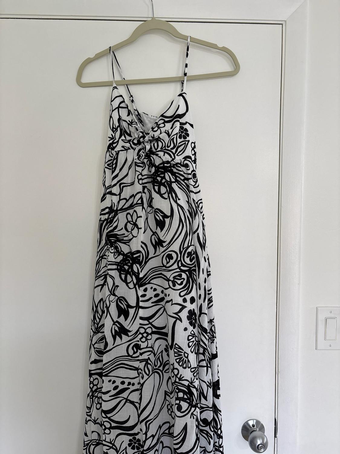 Long dress with thin straps, white with a black botanical print, hanging on a hanger on a white door with a metal knob.