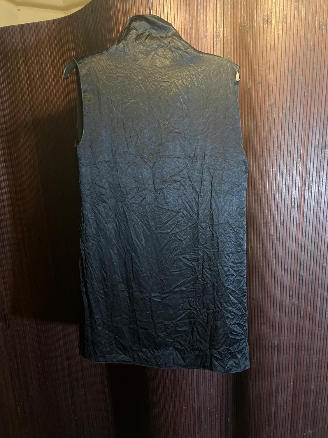 Sleeveless black vest hanging on a hanger, with wrinkled texture, against a brown wood panel background.