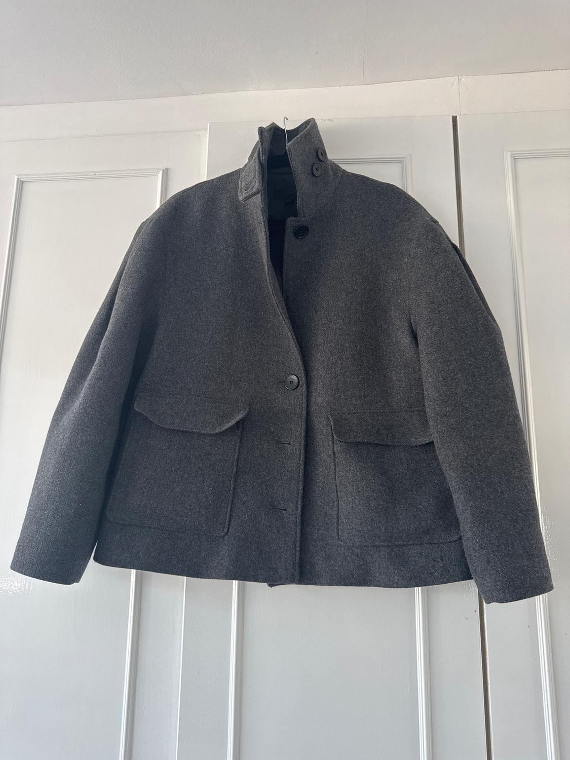 Grey wool jacket hanging on a white door, with a high collar and two large pockets on the front.