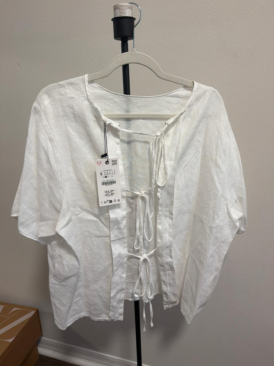 Short-sleeve white blouse with front ties, hanging on a hanger. Visible tag with size and price details.