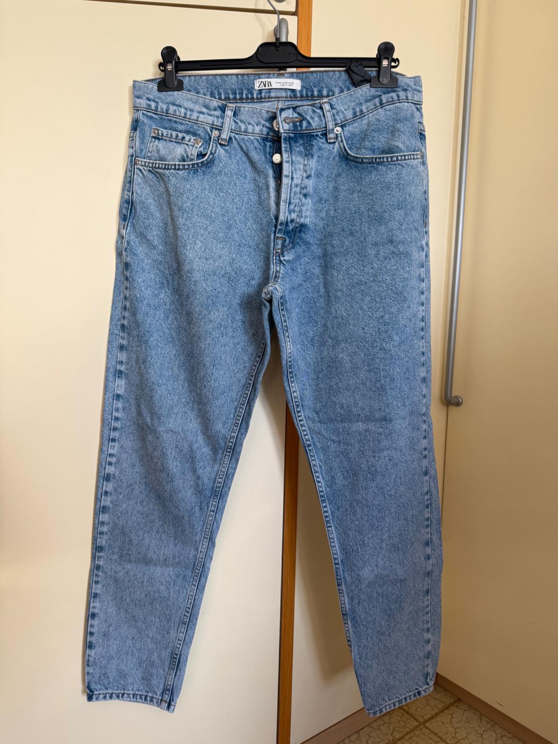 Light blue jeans hanging on a black hanger against a beige door. Visible label with the text "ZARA".