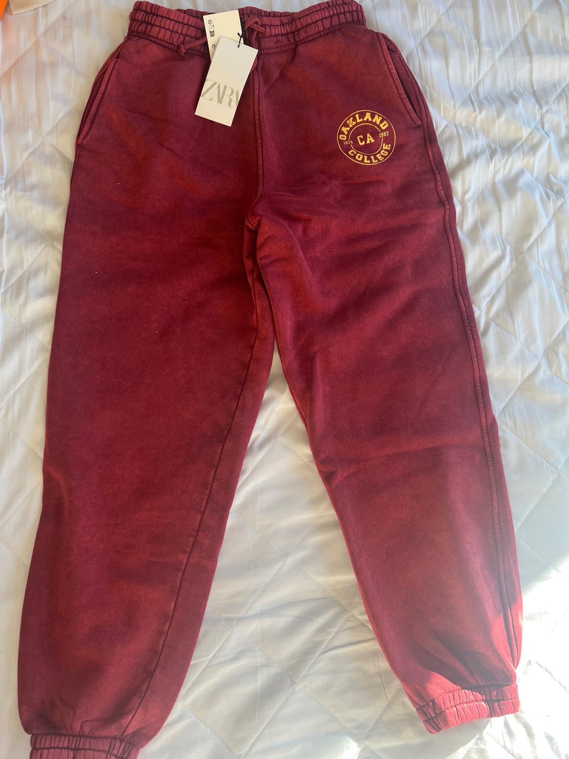 Burgundy jogger bottoms with elasticated waist and ankles, "Oakland College CA" logo on the thigh, on a white bed.