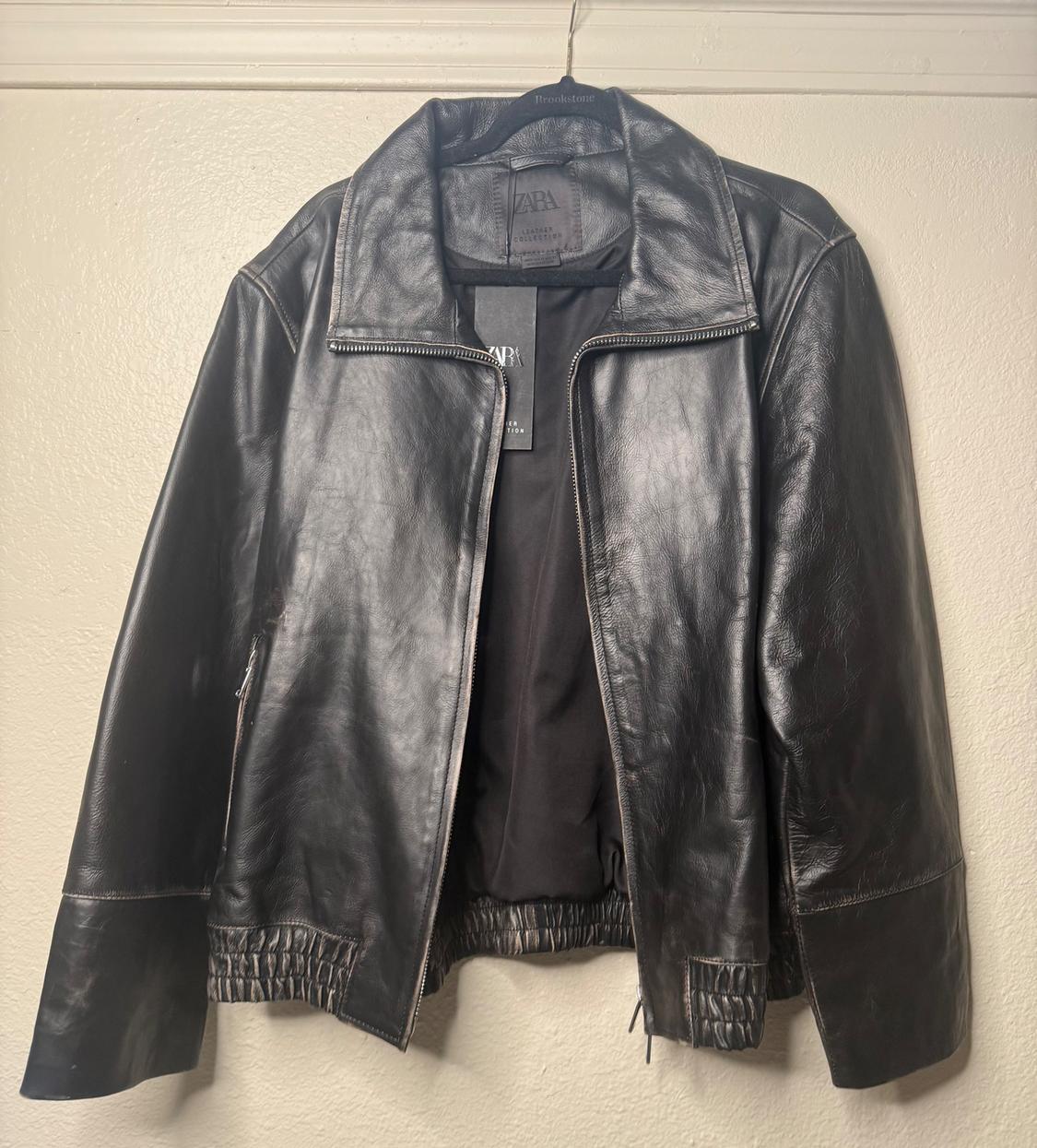Black leather jacket hanging on a hanger, with a front zipper and visible Zara tag. Beige wall background.