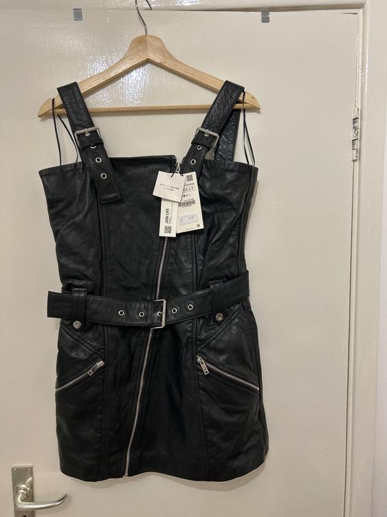 Black dress with wide straps, front zip, and belt, hanging on a wooden hanger over a white door.