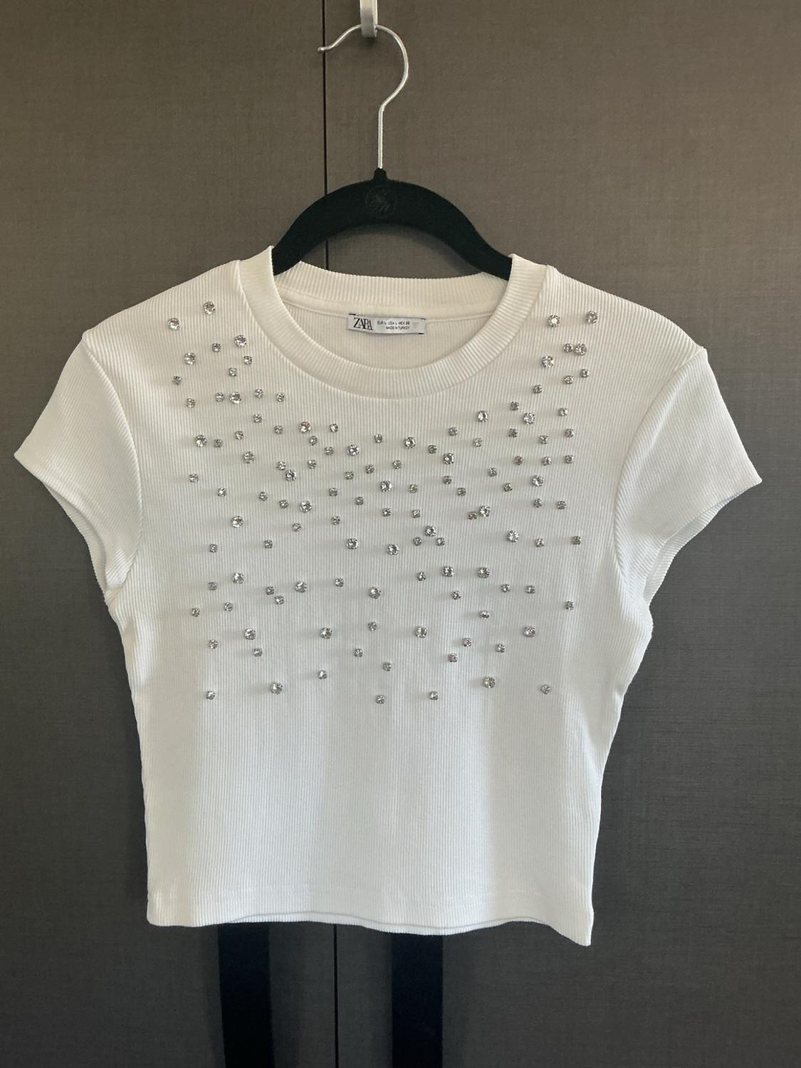 White ribbed short-sleeve t-shirt with shiny rhinestones, hanging on a black hanger against a dark background. ZARA label.