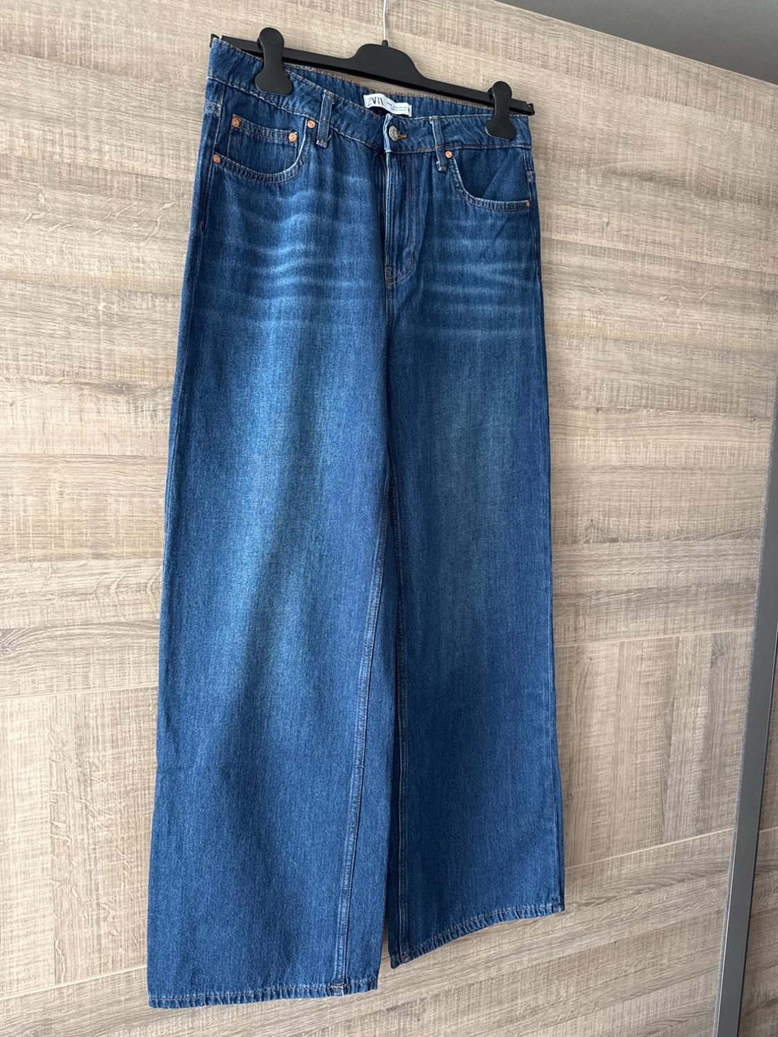 Baggy blue denim jeans hanging on a black hanger against a light wooden background. Label visible on the waistband.