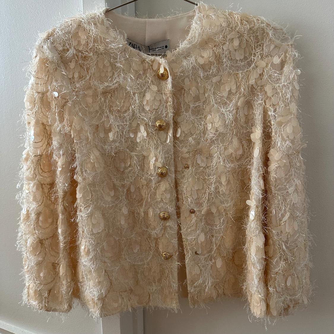 Beige jacket with sequin texture and gold buttons, hanging on a hanger against a white background.