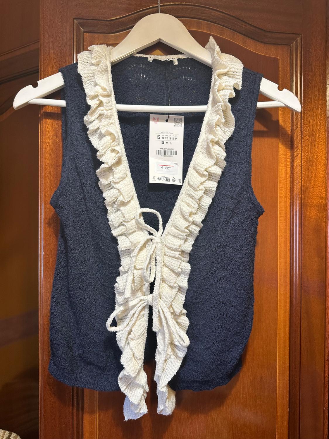 Navy knitted waistcoat with white ruffles and bows, on a wooden door. Text: S S 26 S S P € 22.95.