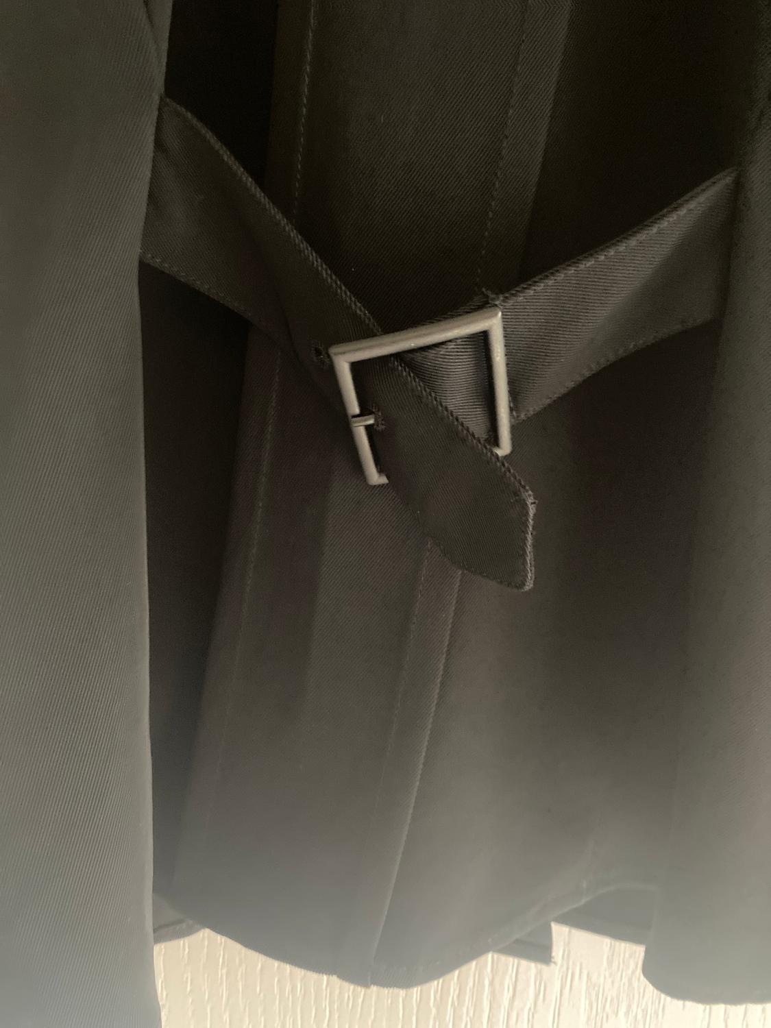 Detail of a black coat with a belt and square metal buckle, hanging on a light-coloured wall.