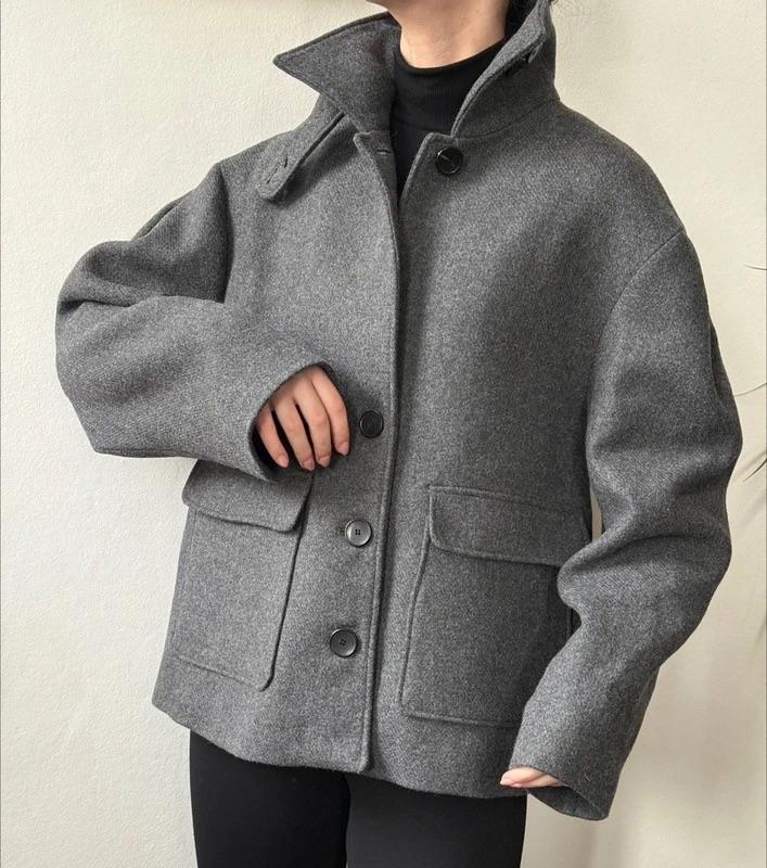Model wearing a grey wool jacket with large buttons and a high neck, paired with a black jumper. Minimalist white background.