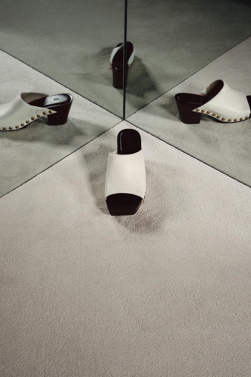Front view of a white sandal with a dark heel, an open square toe, and metal studs on the sides.