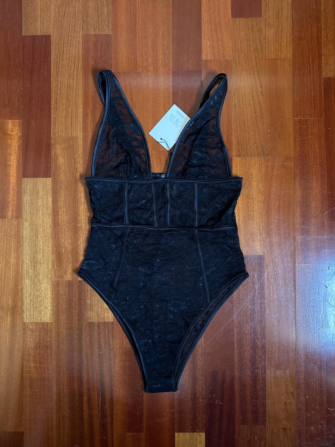 Black floral lace bodysuit with a deep V-neckline and seam detailing, on a wooden floor. Label with the text ZARA LINGERIE.