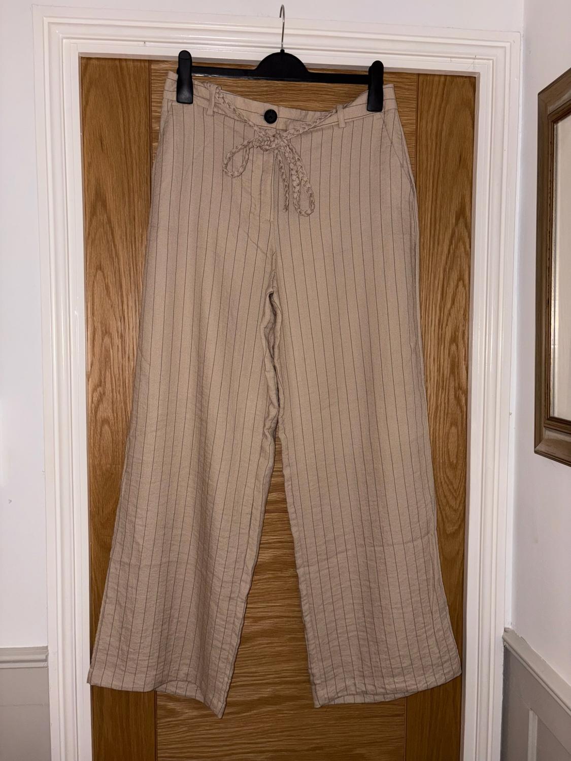 Beige wide-leg striped trousers with a braided belt, hanging on a coat hook over a wooden door.