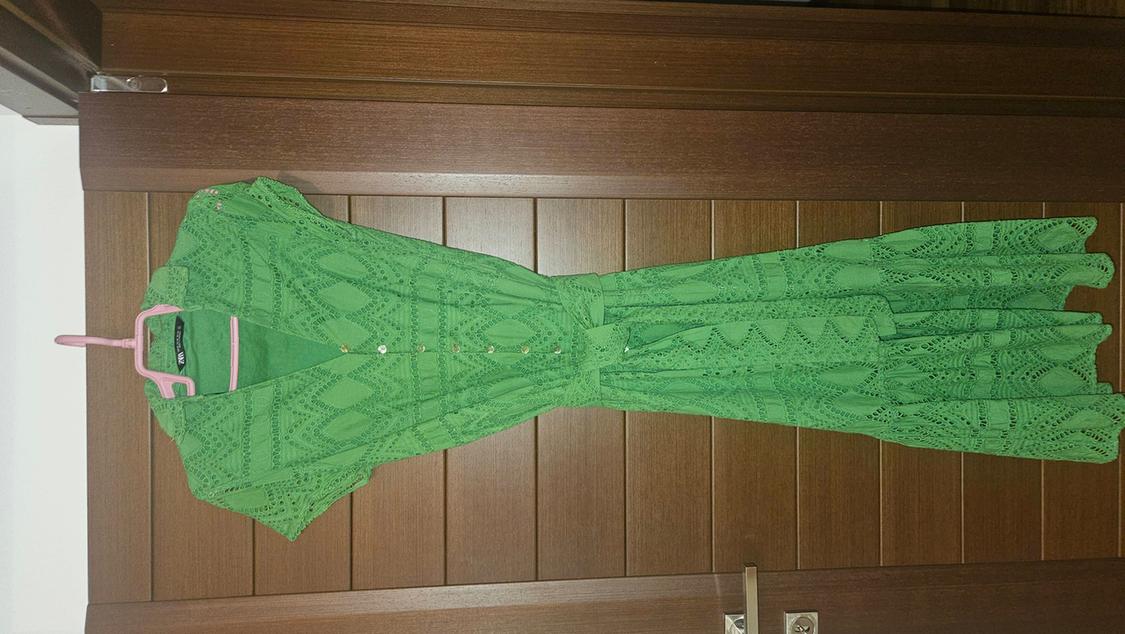 Green lace dress hanging on a pink hanger over a wooden door. Geometric pattern design with front buttons.