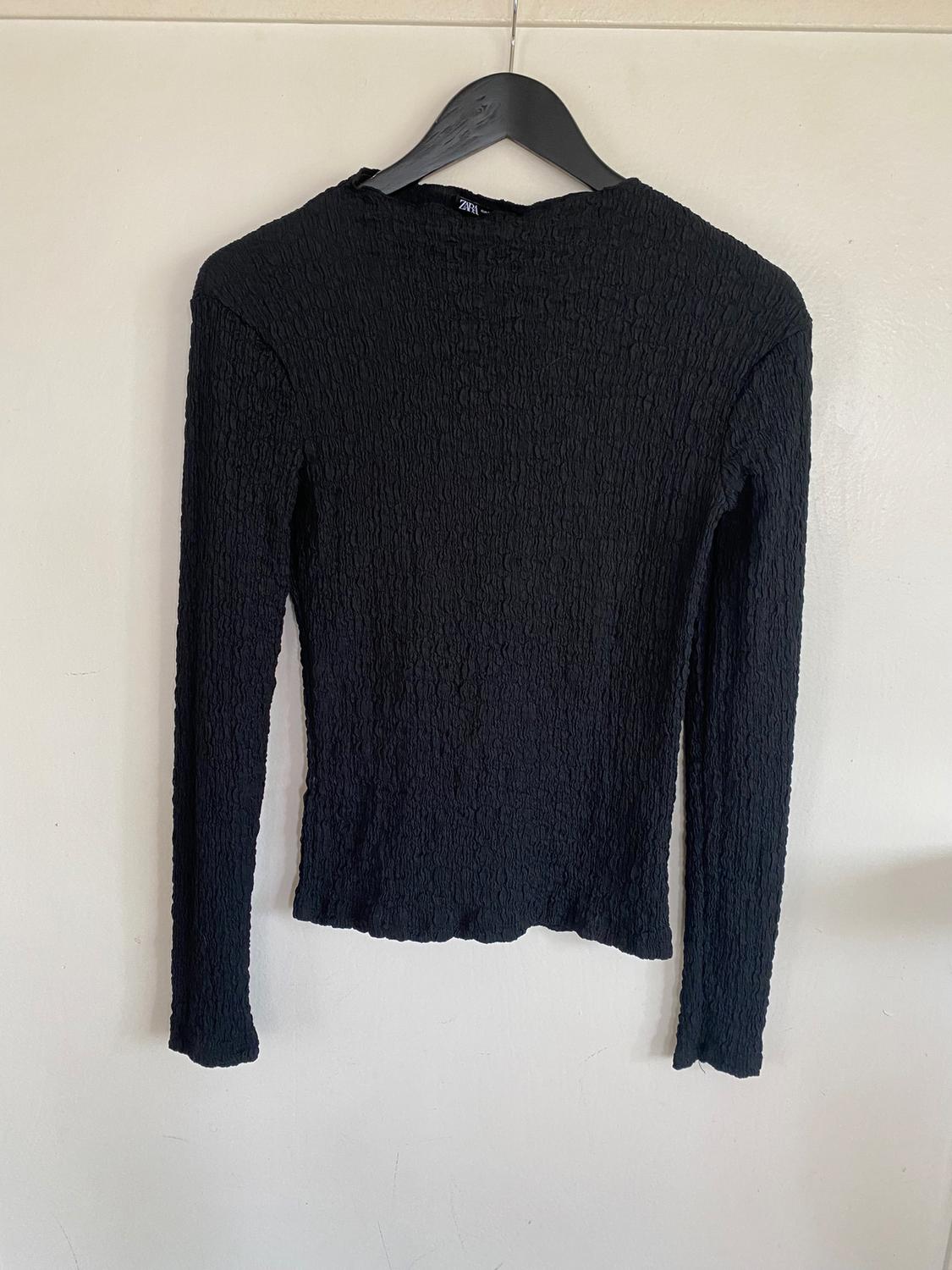 Black long-sleeved textured crinkle-effect jersey, hanging on a black hanger against a white wall.