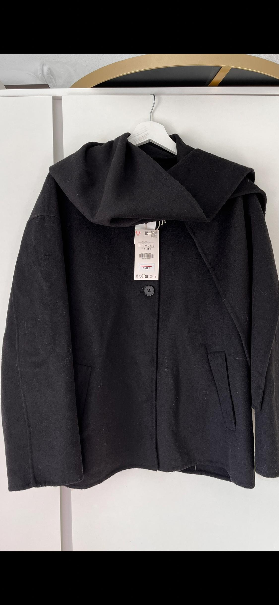 Black coat with a hood hanging on a white hanger. Visible label with text and a barcode. Black and white background.