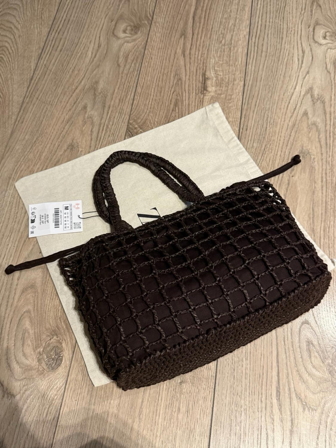 Dark brown woven net-style tote bag over a fabric bag with the letter Z on a wooden floor. A label showing size M can be seen.