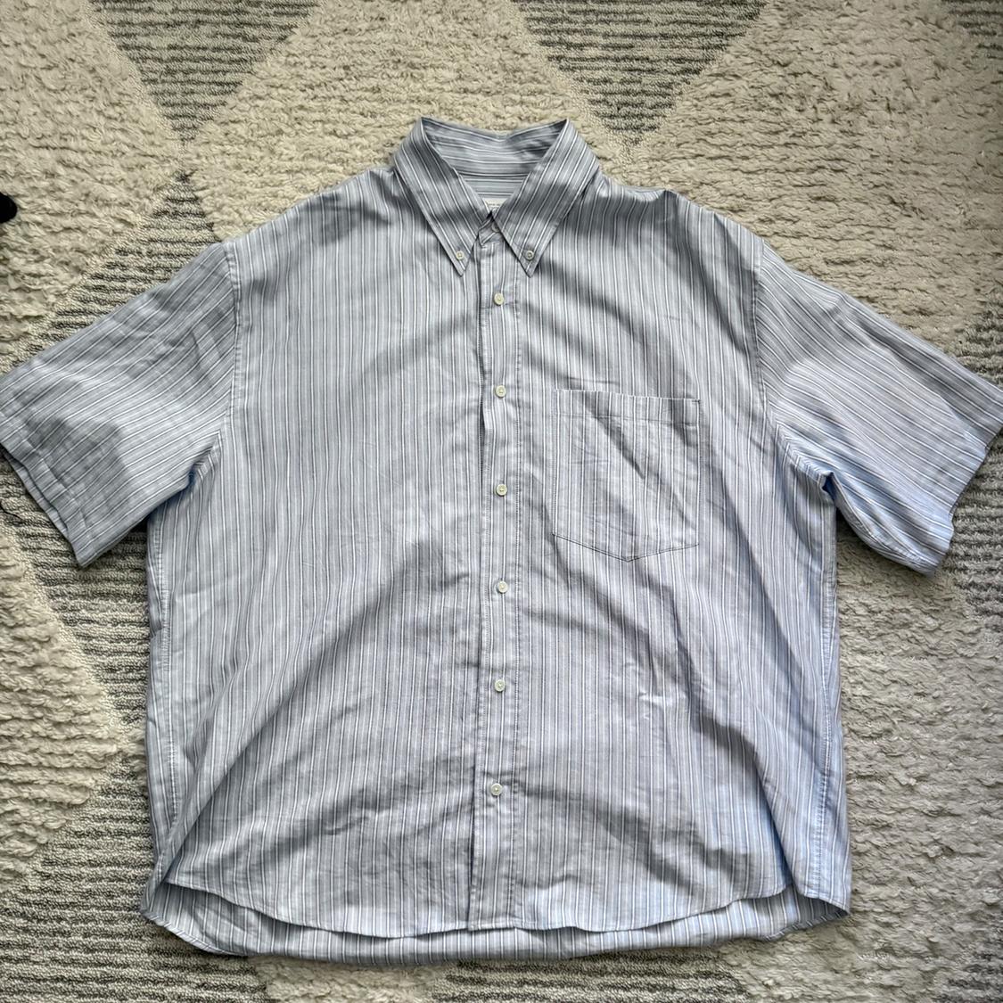 Blue and white striped button-down shirt, short-sleeved with a chest pocket, on a soft-textured rug.