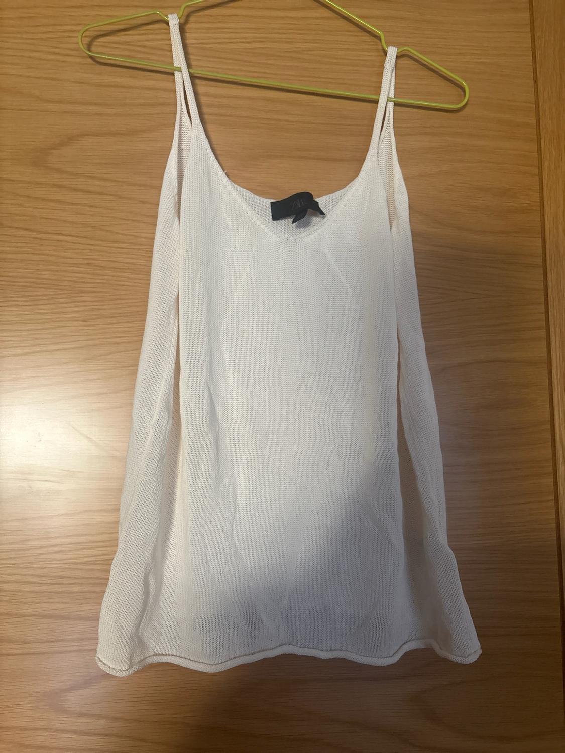 White fine-knit V-neck top with double straps, hanging on a yellow hanger against a wooden background.