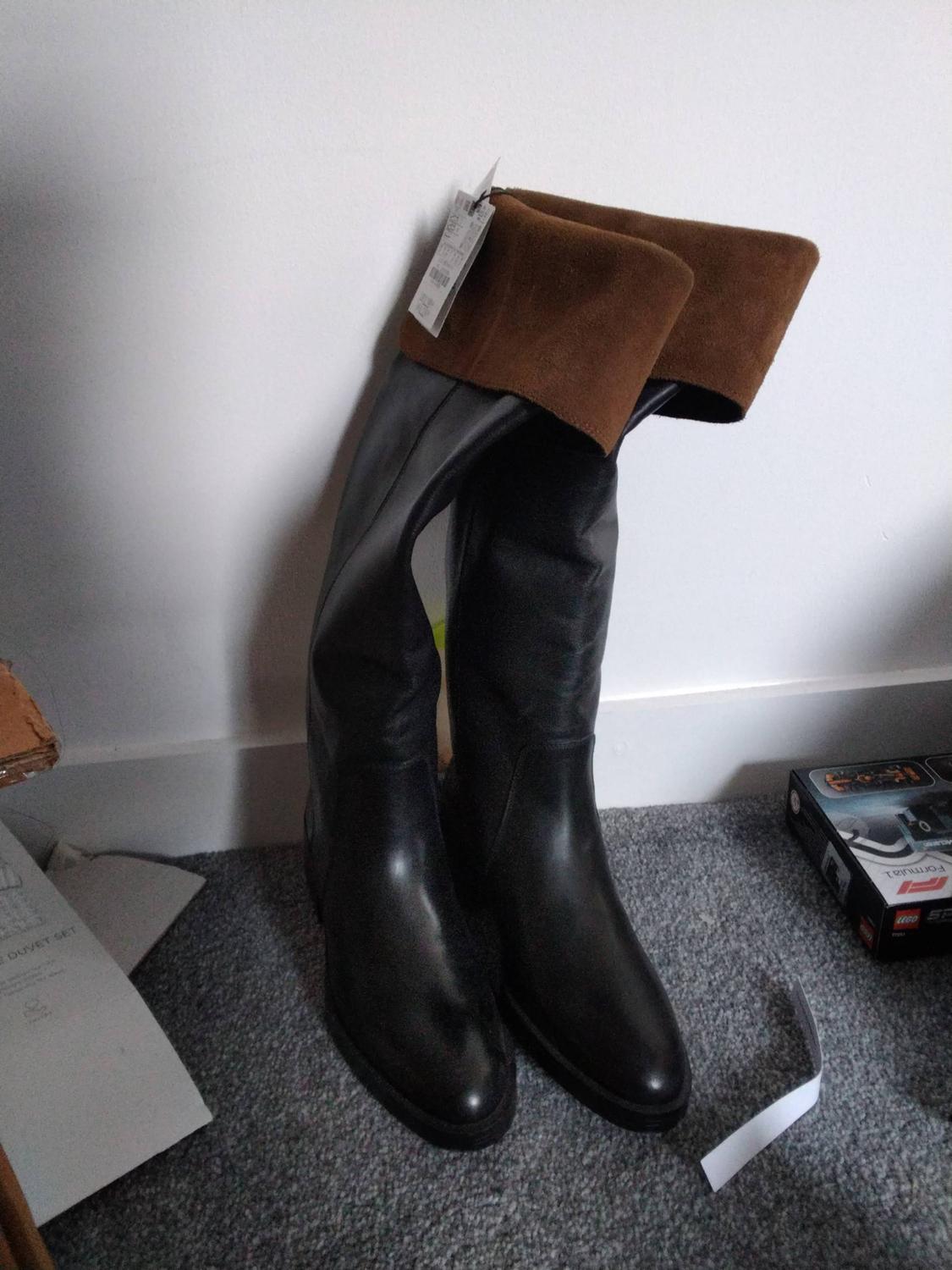 Black high boots with tags and a brown turn-down, leaning against a white wall on a grey carpet. Texts: DUVET SET, LEGO, Formula 1.