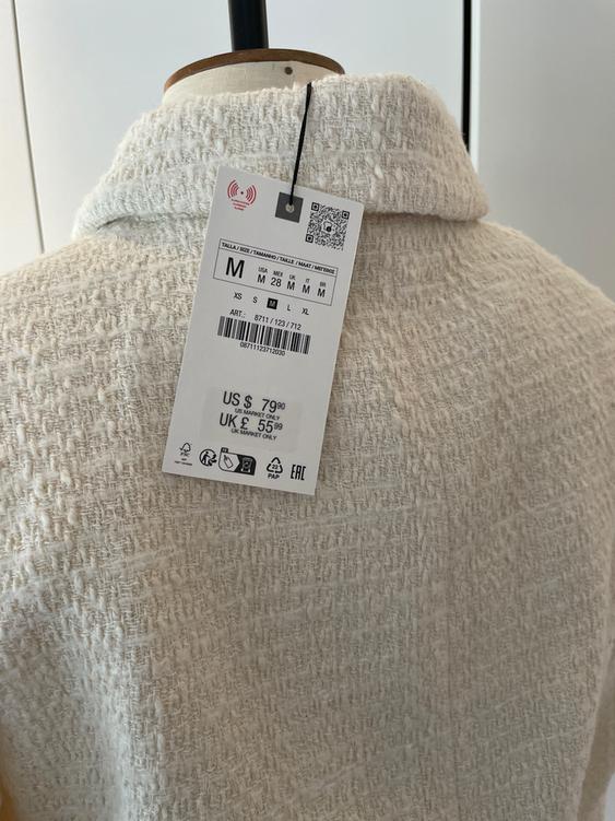 Price tag on a white textured fabric coat. Price: US $79.90, UK £55.99. Size M. Barcode visible.