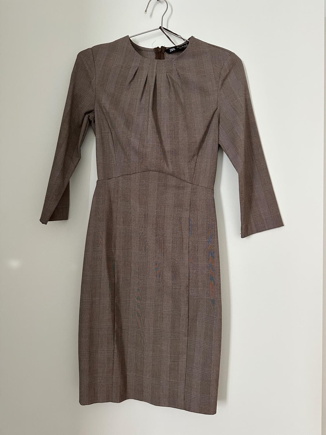 Brown long-sleeved dress hanging on a white hanger. Simple design with pleats on the chest and a back closure.