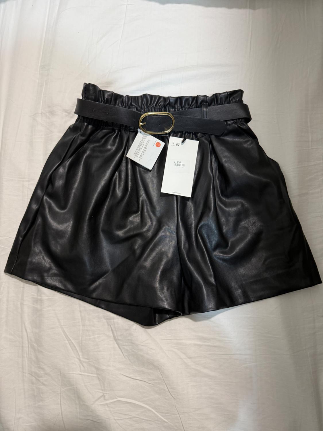 Black shiny material shorts with a belt and gold buckle, on a white surface, with hanging tags.