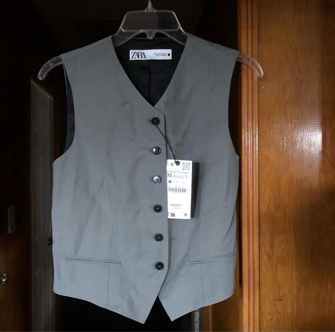 Gray sleeveless vest hanging on a clear plastic hanger, with a visible price tag, in a wooden setting.