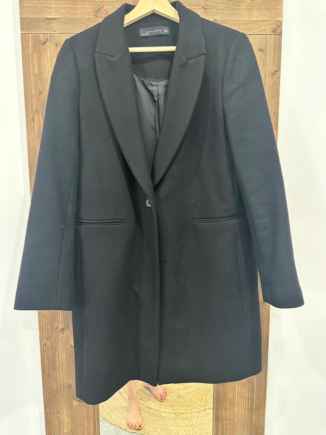 Black coat with a ZARA WOMAN label on a wooden hanger. Below, a mirror reflects bare feet with red toenails on a rug.