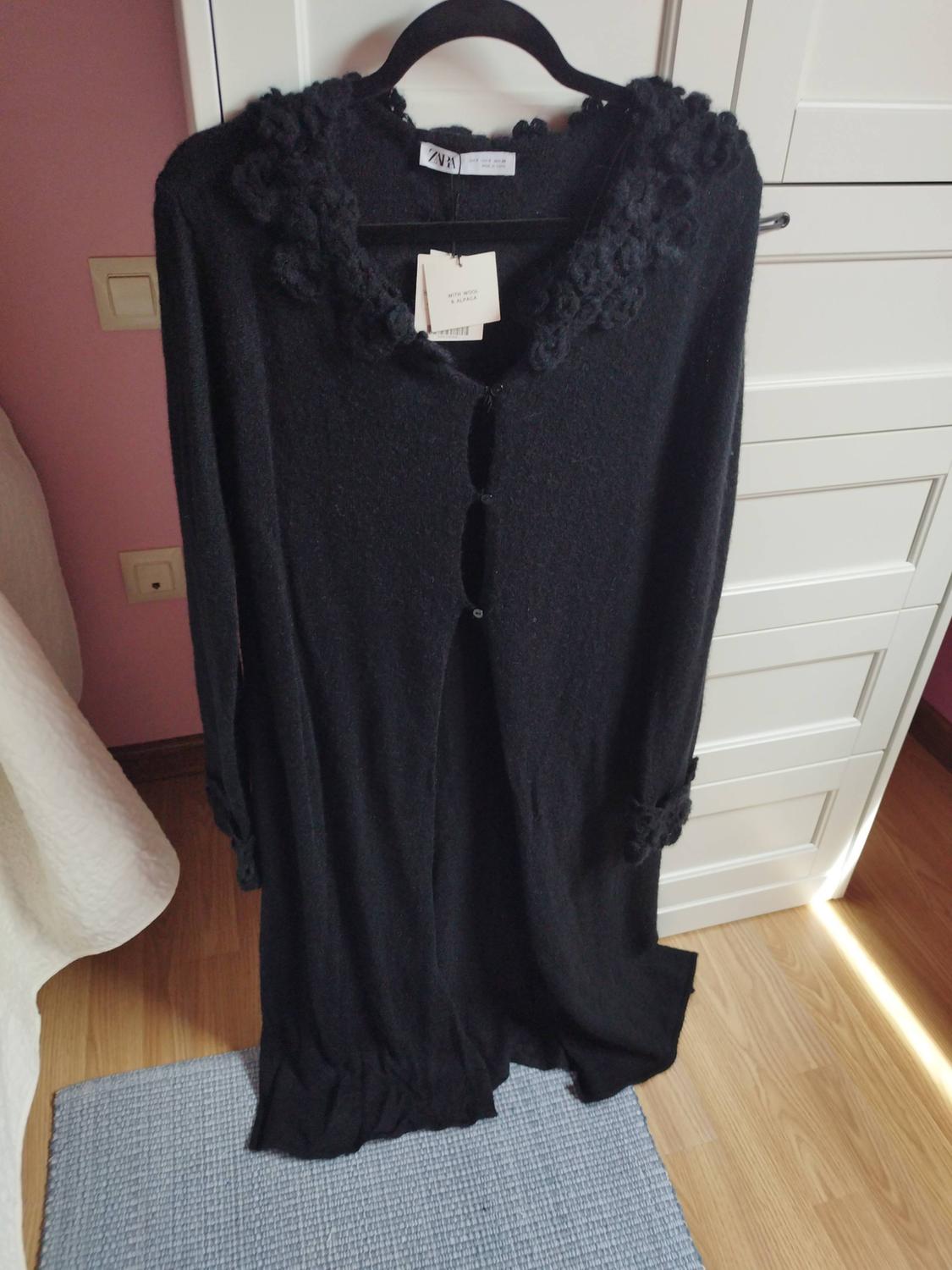 Long black button-up cardigan with collar and cuff detailing, hanging in a room with a pink wall and wooden floor. Text: ZARA.