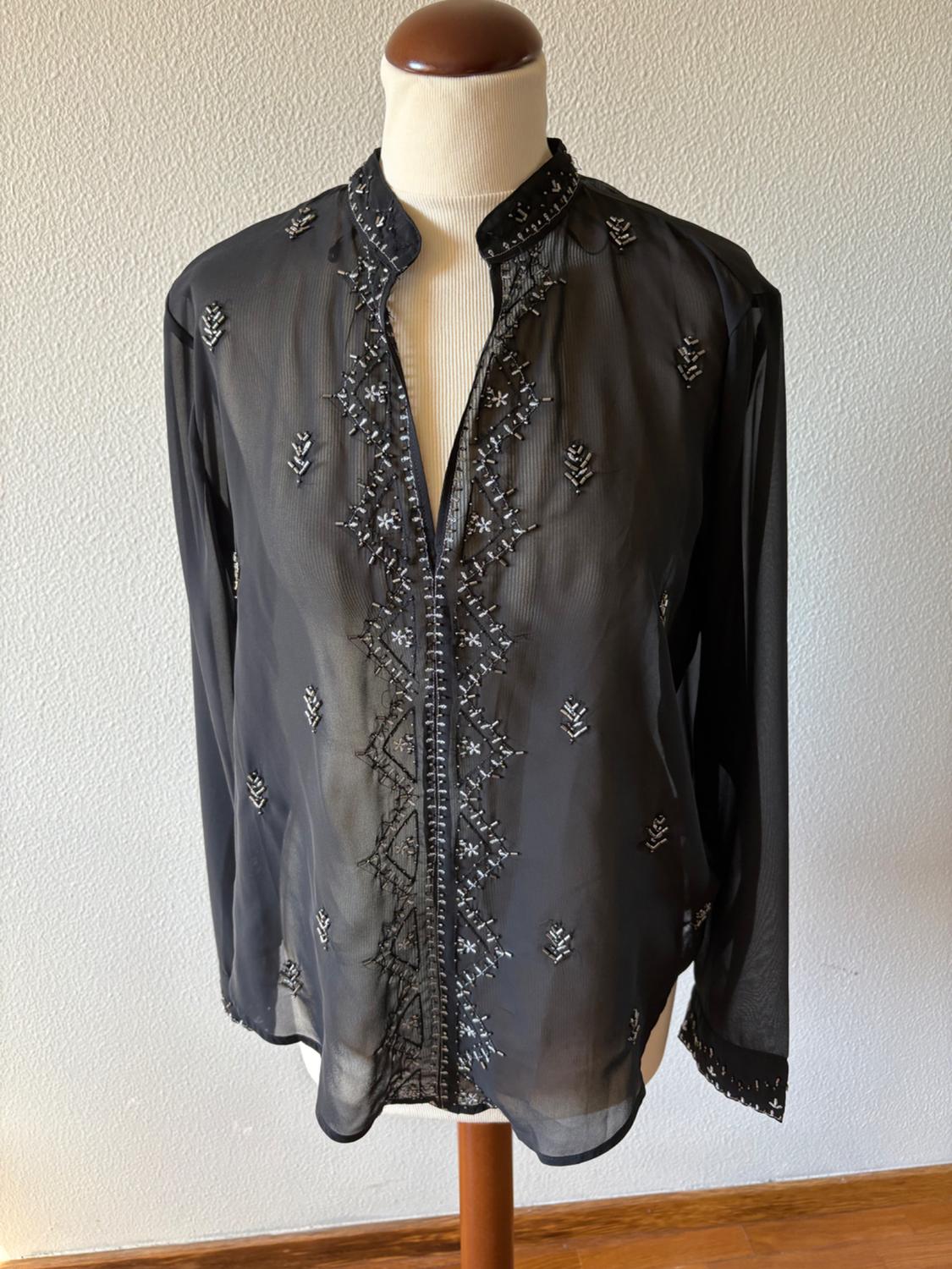 Black semi-transparent long-sleeved blouse with silver embellishments, displayed on a mannequin against a white wall background and wooden floor.
