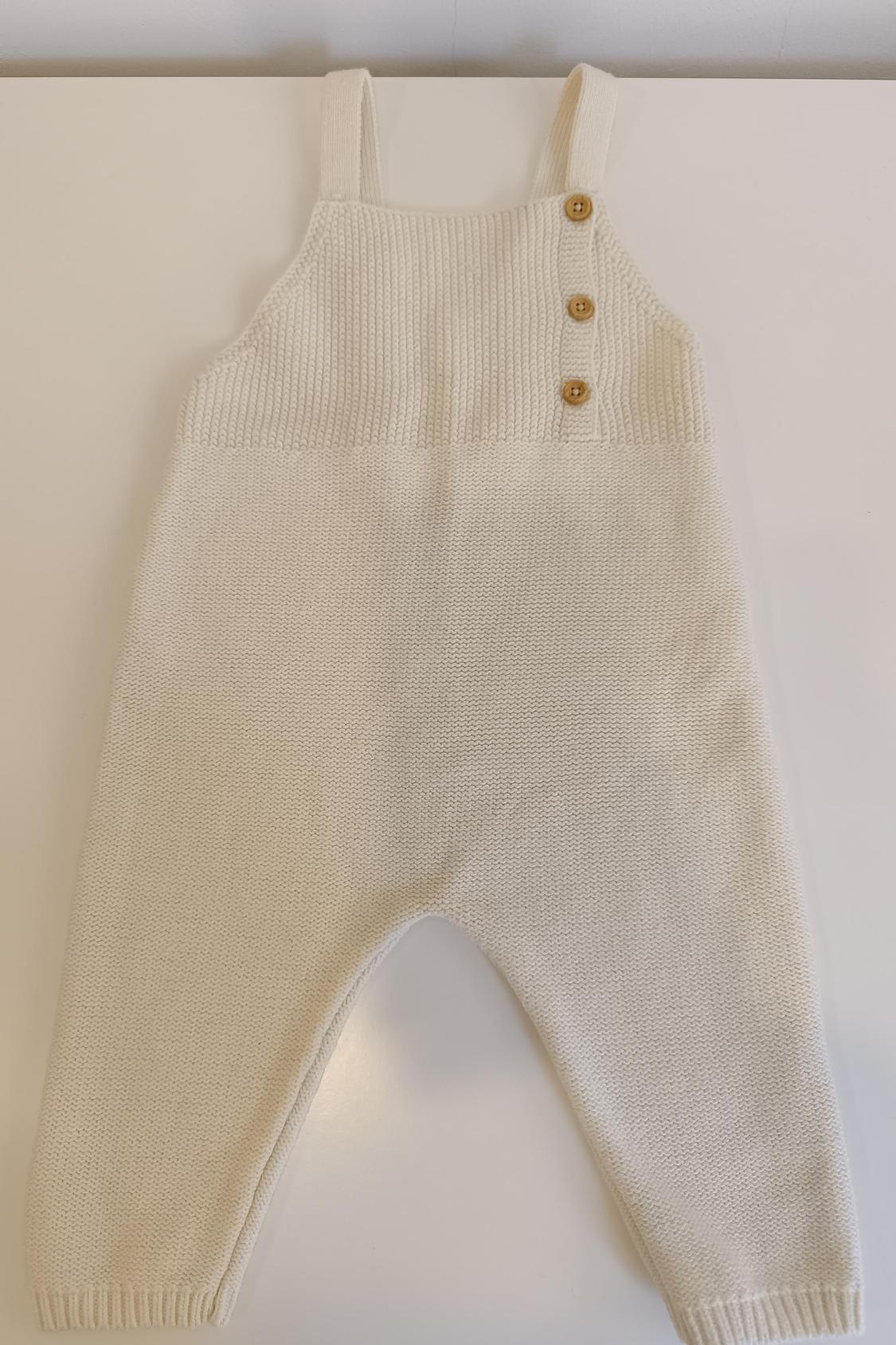 Front view of a beige knit jumpsuit with straps and wooden buttons on the chest.