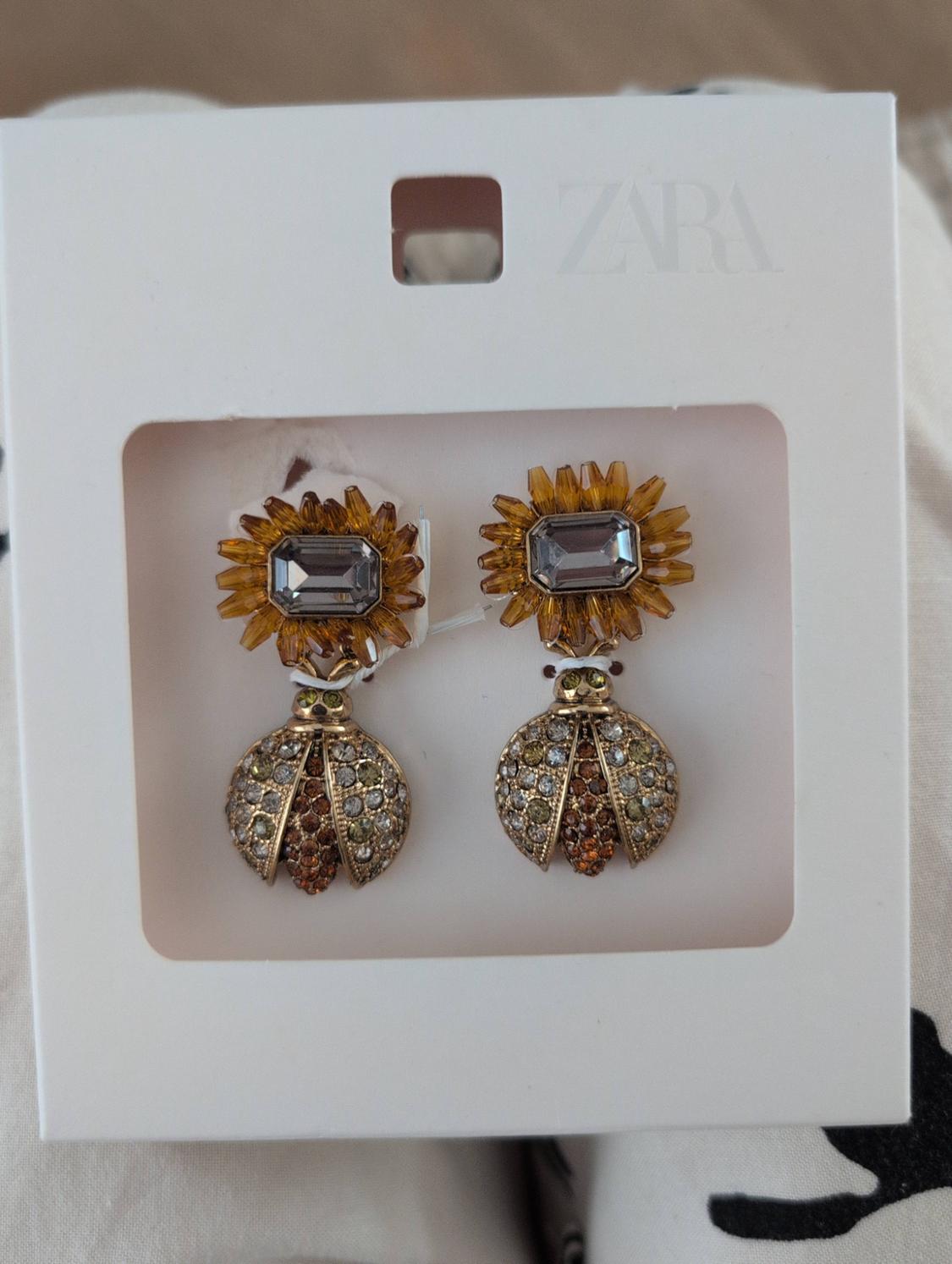 White cardboard earrings with ZARA text. Flower design with amber petals and a grey centre, with a hanging insect featuring sparkling stones.