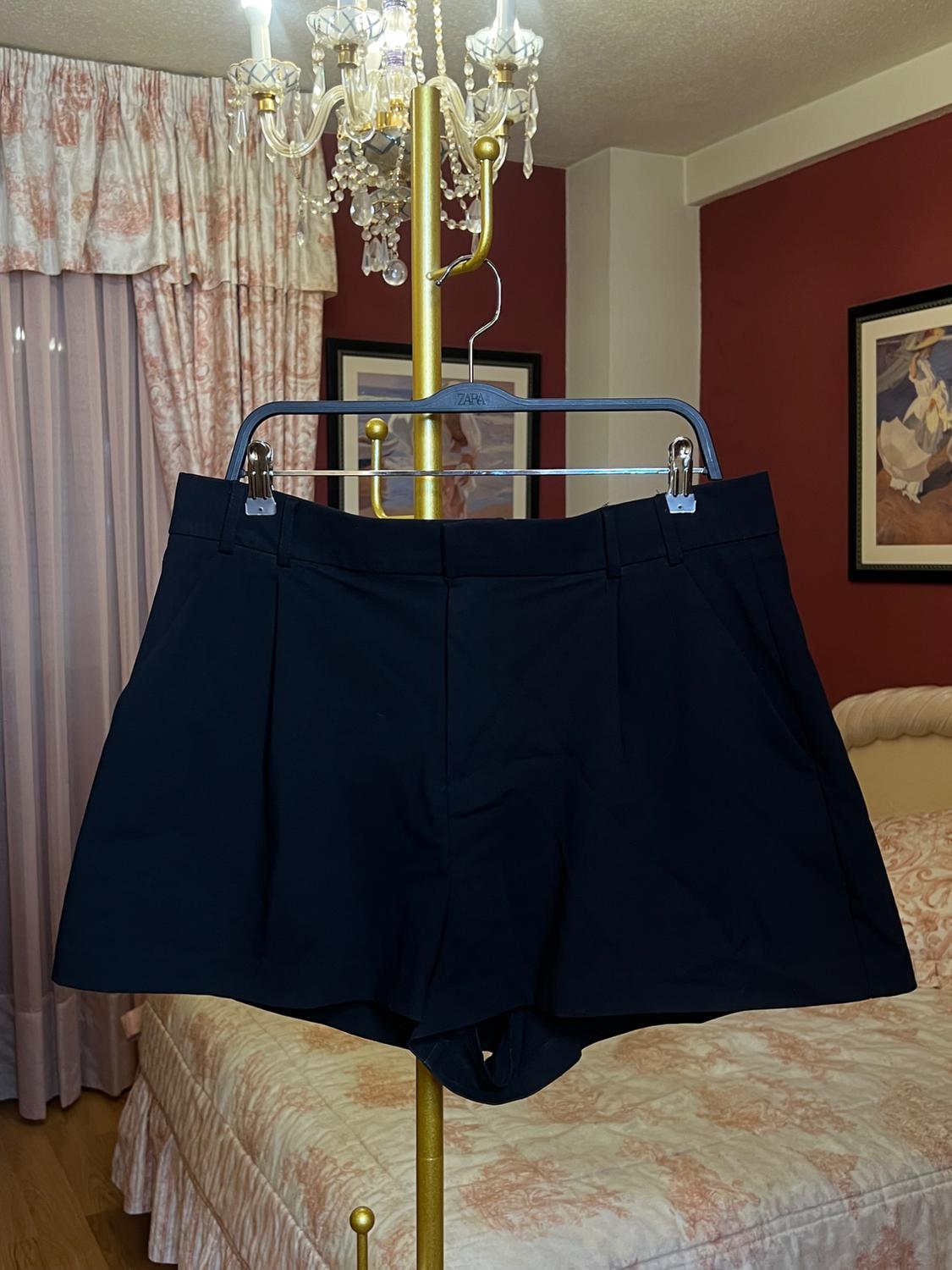 Black shorts hanging on a gold lamp in a bedroom with red curtains and walls. Hanger with "ZARA" text.