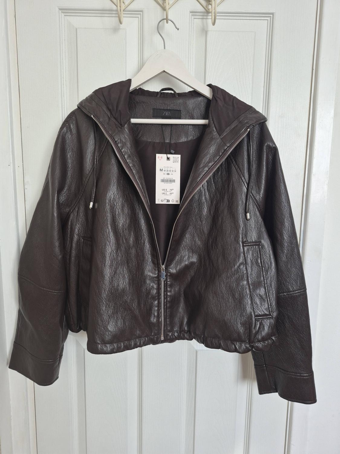 Dark brown jacket with a hood and a zip, hanging on a coat hanger over a white door. Label with text ZARA.