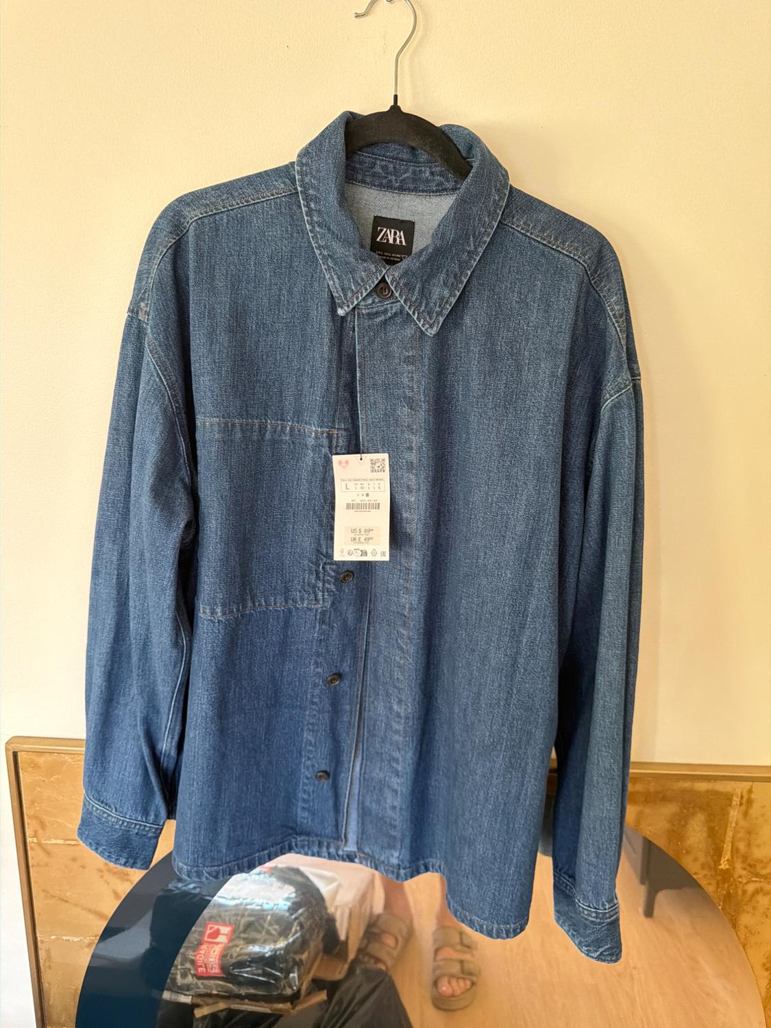 Blue denim shirt hanging on a black hanger with a visible Zara tag. Reflection of feet in sandals in the mirror below.