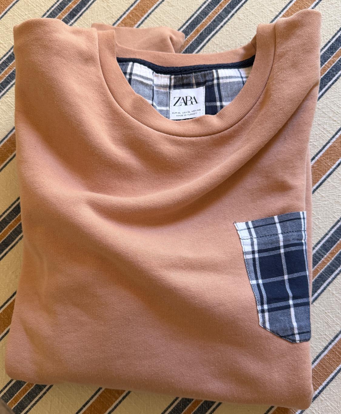 Camel sweatshirt folded over a striped background. Plaid pocket and collar detail. Tag: ZARA EUR XL USA XL MEX 44 MADE IN TURKEY.
