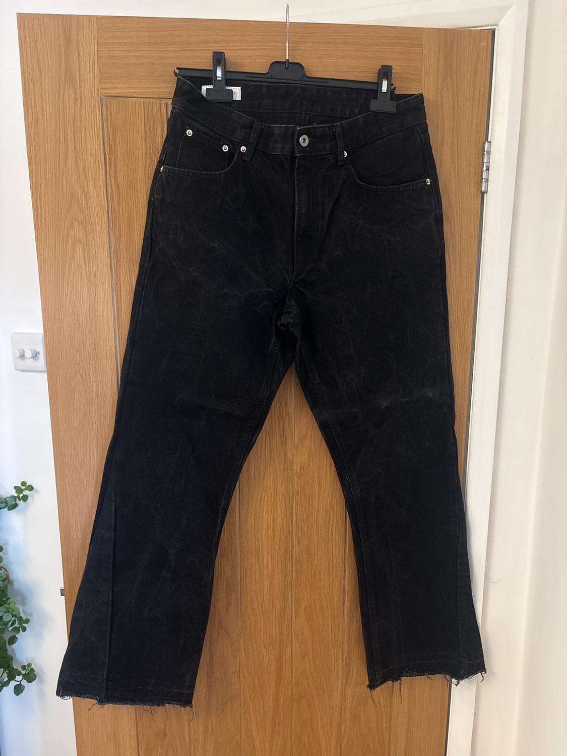 Black jeans with a frayed hem hanging from a coat hanger on a wooden door in a relaxed indoor setting.
