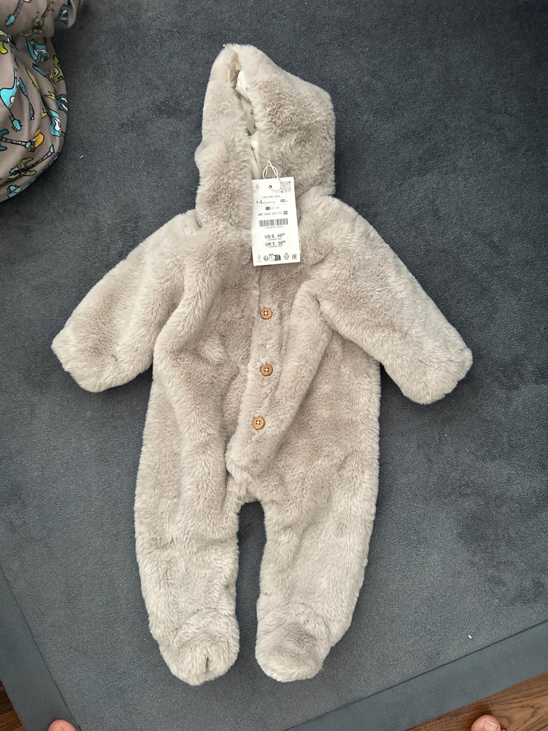 Beige plush baby jumpsuit with hood and front buttons. Visible tag with text US $ 49 UK £ 39. On a gray background.