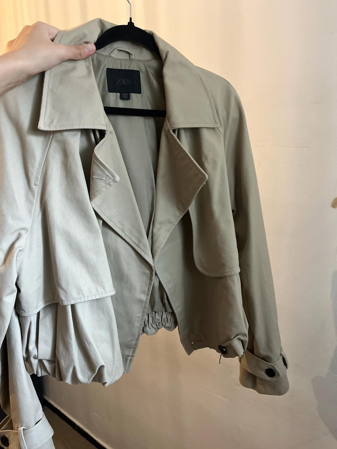 A hand holds a short beige jacket on a black hanger in front of a light-coloured wall. ZARA can be read on the inner label.