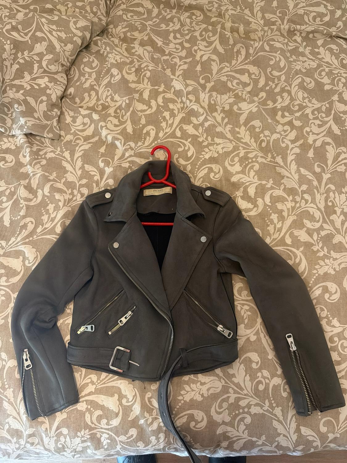 Dark grey biker jacket with zips on a red hanger over a bed with a beige floral bedspread. Text: ZARA BASIC.