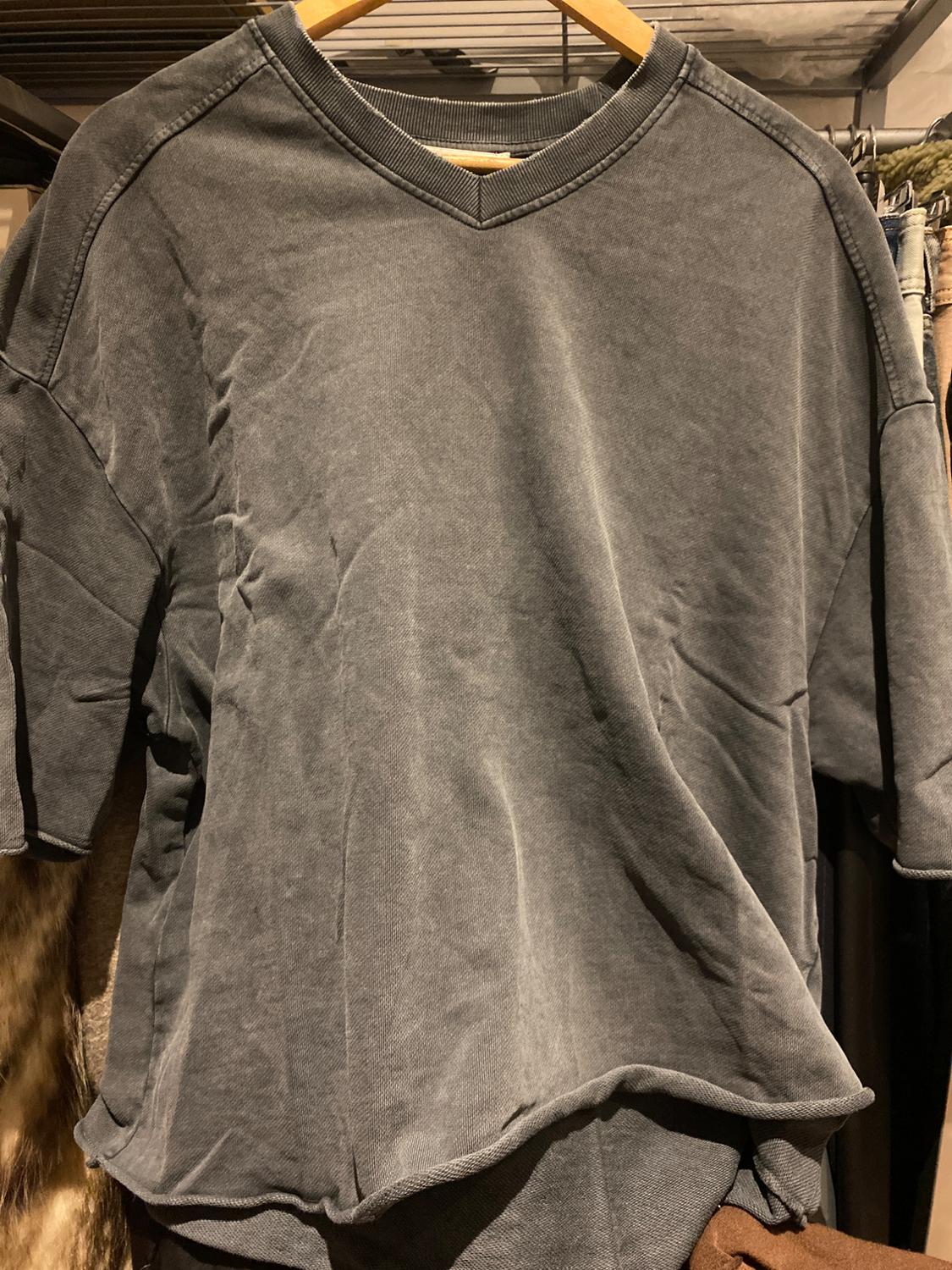 Grey short-sleeved V-neck T-shirt hanging on a wooden hanger, with clothes in the background in a wardrobe.