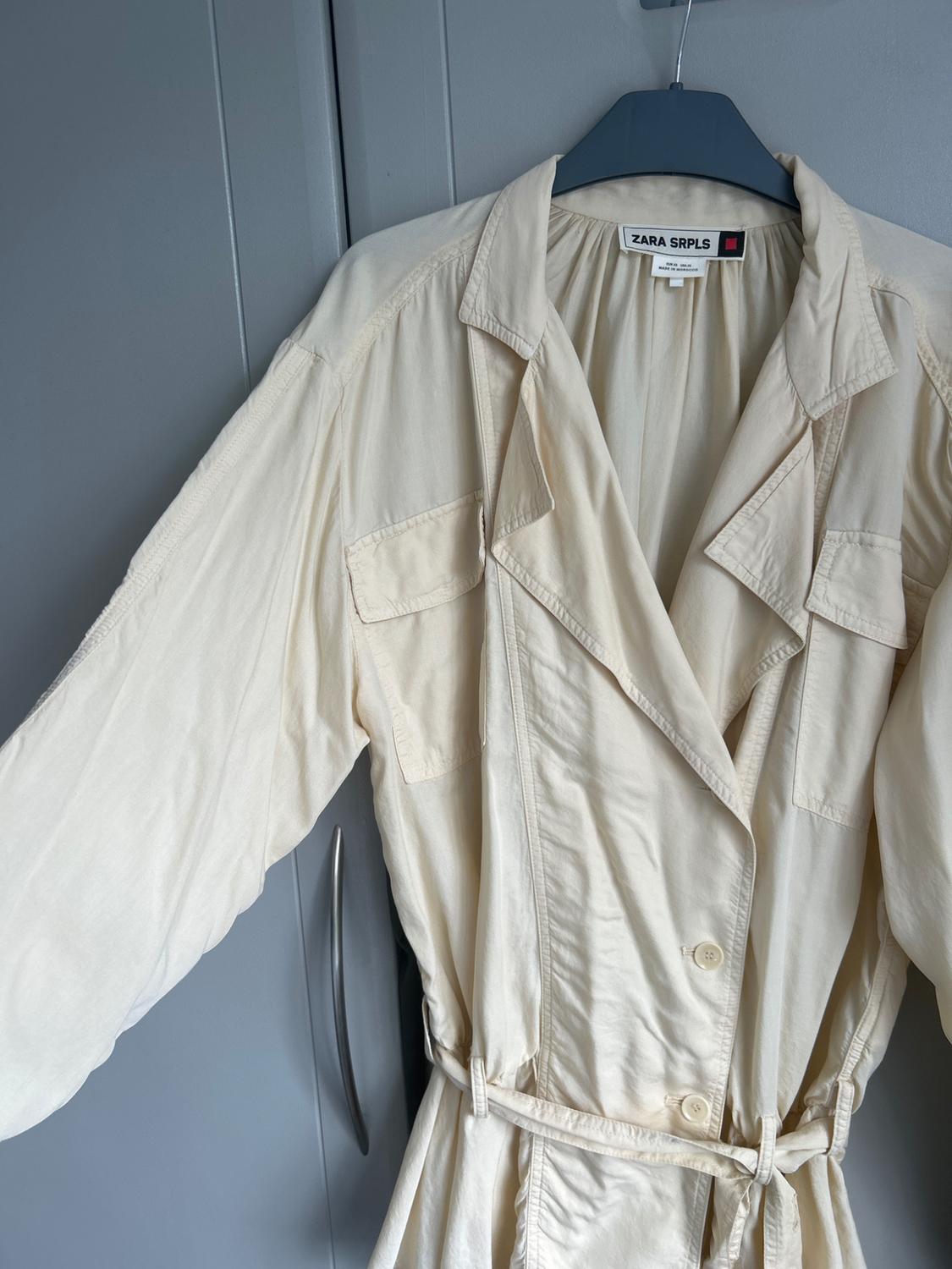 Cream-coloured garment with lapels, pockets, buttons and a belt, hanging on a hanger in front of a grey wardrobe. Text: ZARA SRPLS.