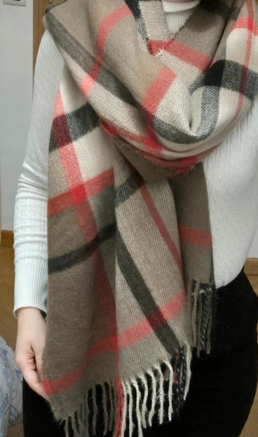 Woman in a white jumper and a checked scarf in shades of beige, red, and black, with fringes. Wooden interior background and white wall.