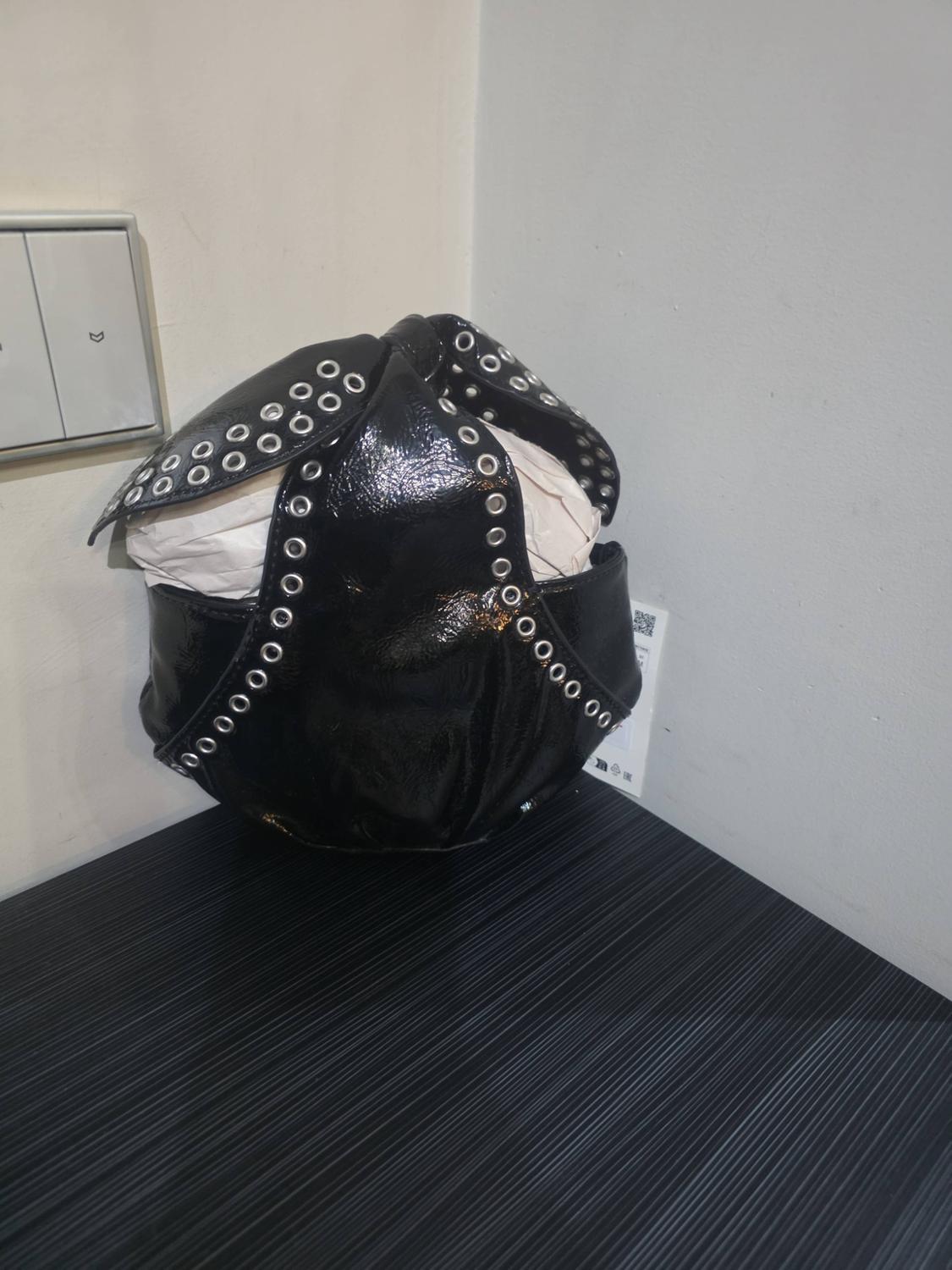 Shiny black handbag with silver metal rings, stuffed with white paper, on a dark striped surface in front of a white wall.